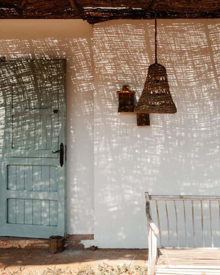 Helle Terrasse auf Ibiza mit blauer Holztür, geflochtener Hängelampe, einer Bank aus Stöcken und lebhaften Lichtmustern an der Wand