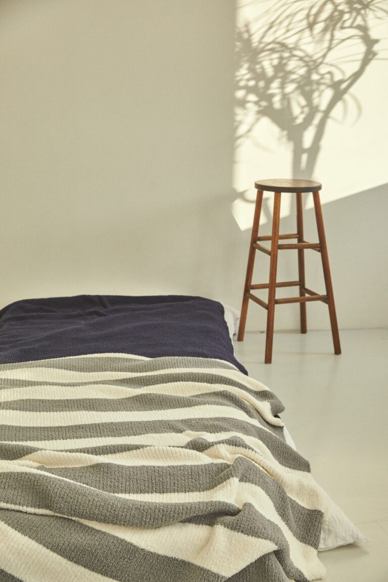 Schlichtes Schlafzimmer mit Bett, gestreifter Decke und Holzstuhl im warmen Sonnenlicht
